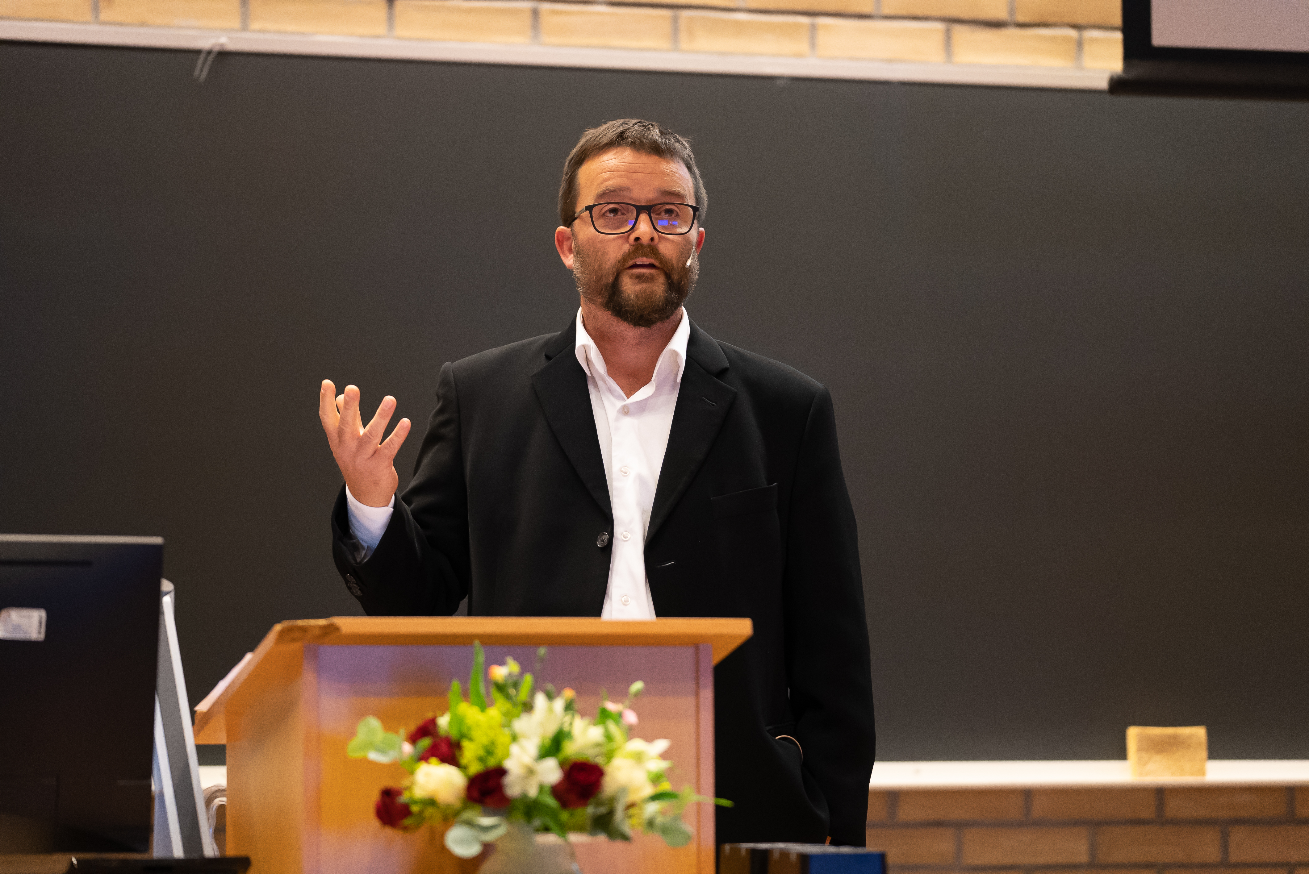 Mann i dress med briller som holder opp en hånd under en tale