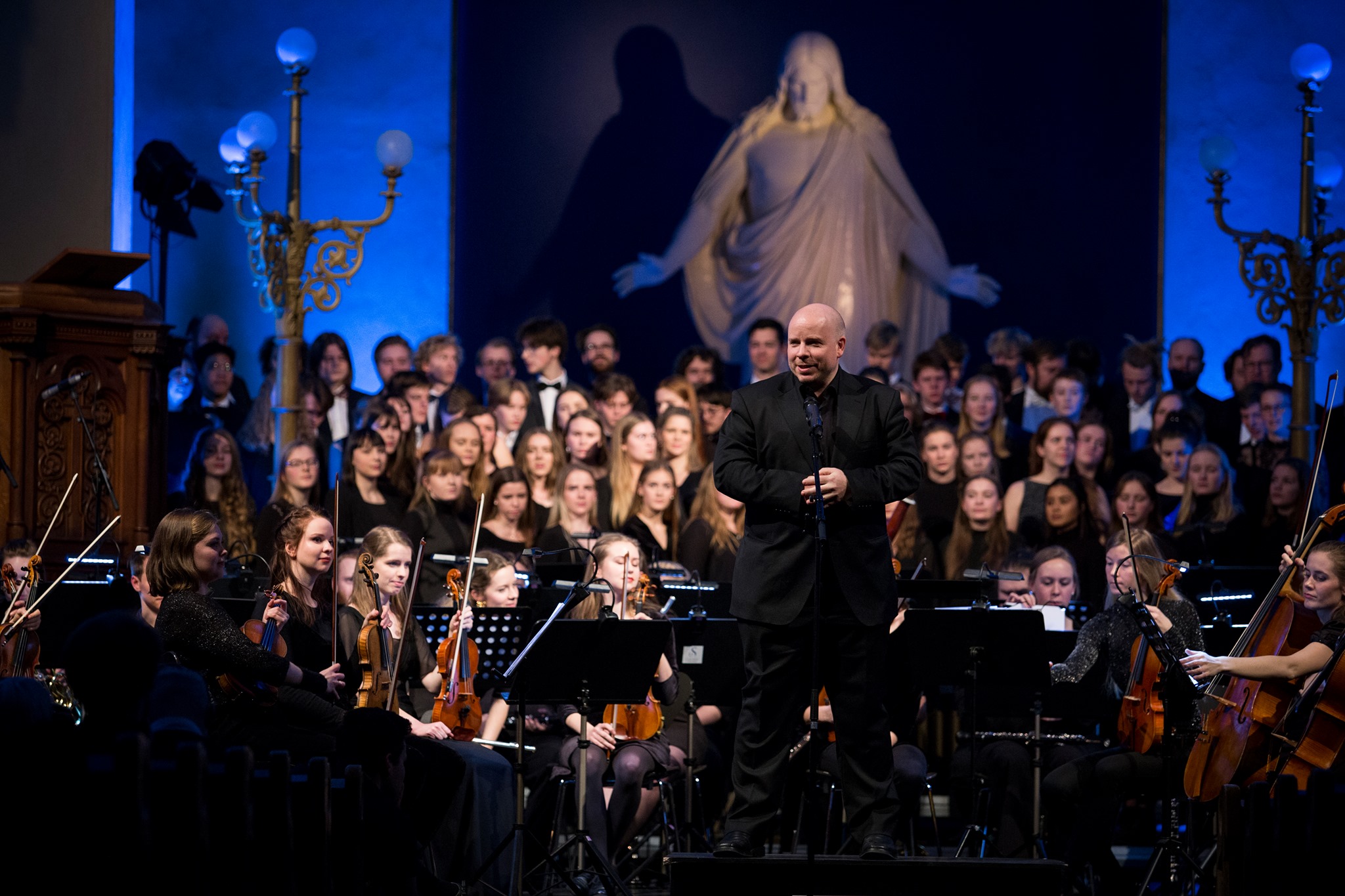 Smilende dirigent foran et stort orkester i en kirke. Fotografi.