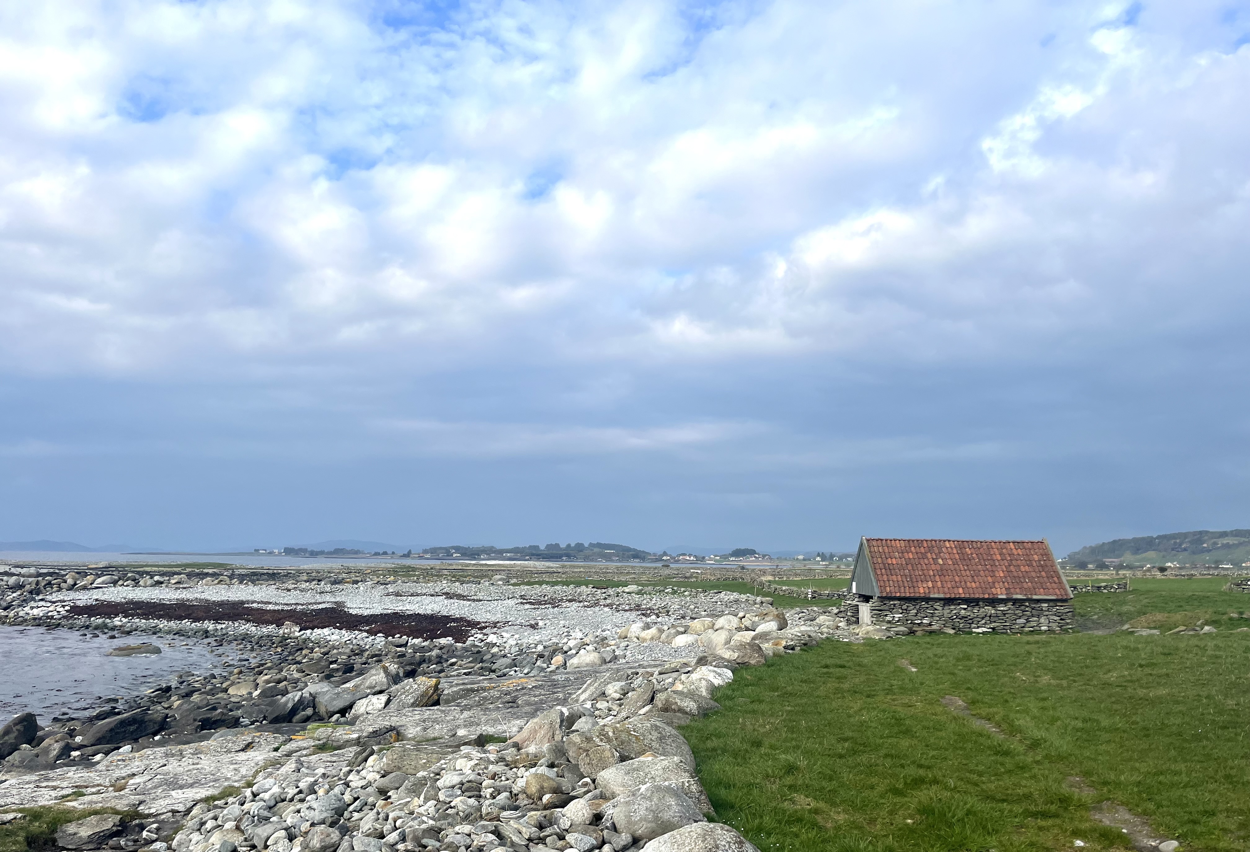 rullesteinstrand, himmel med skyer, grønn utmark, et naust