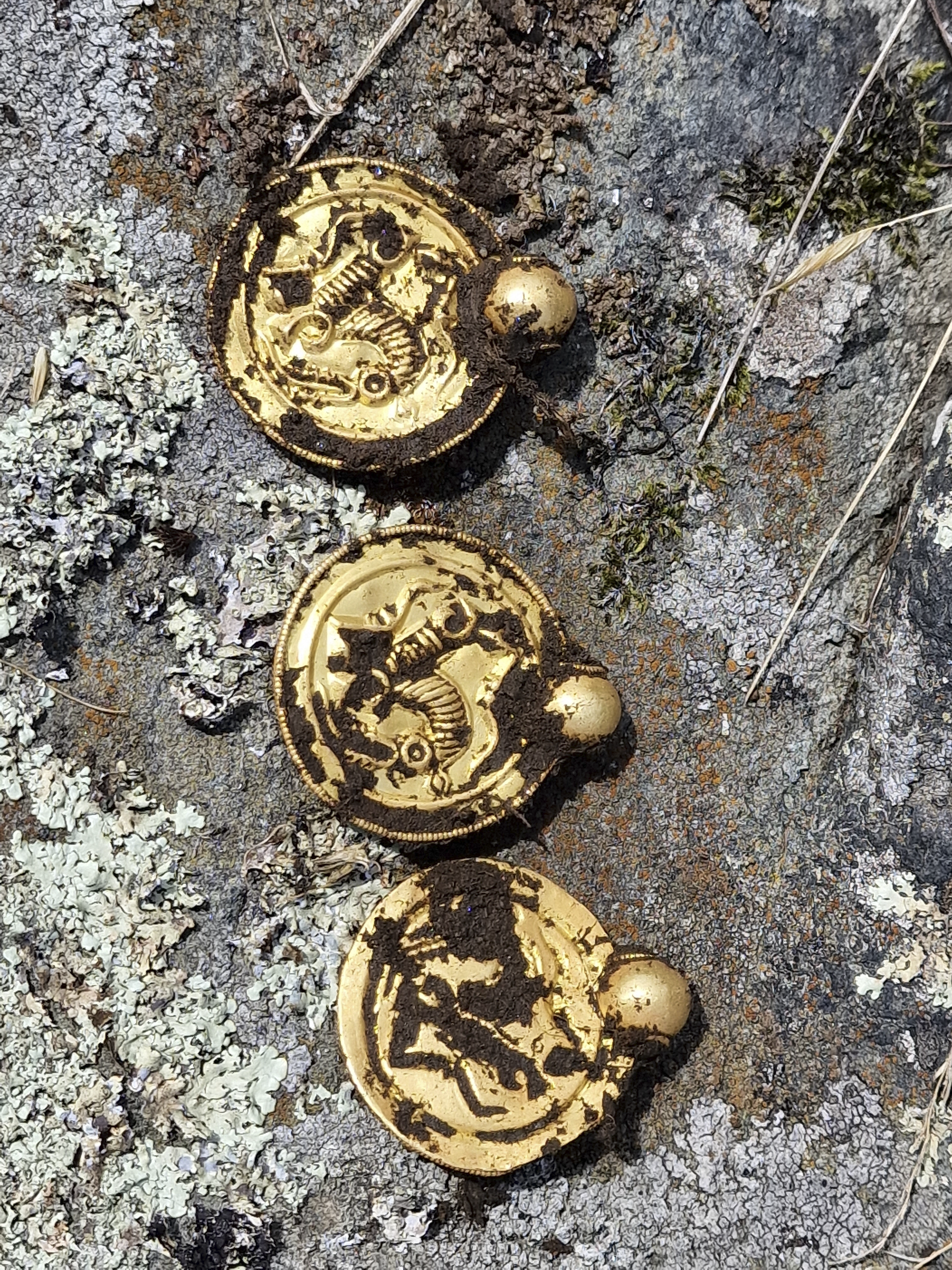 Tre gullmedaljonger - gullbrakteater - med hver sin gullperle liggende i jorden