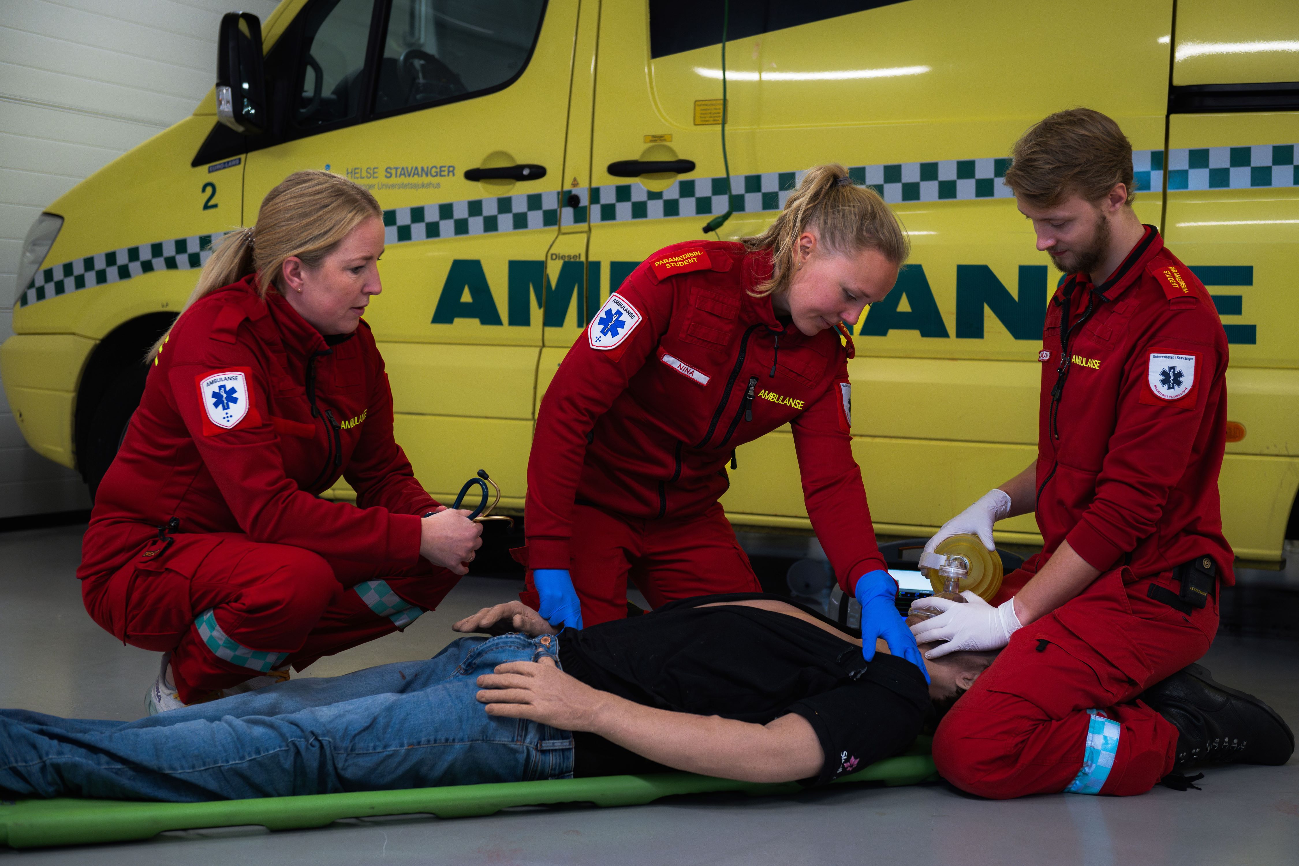 Tre personer i røde ambulanseuniformer trener livredning på en dukke foran en gul ambulanse.