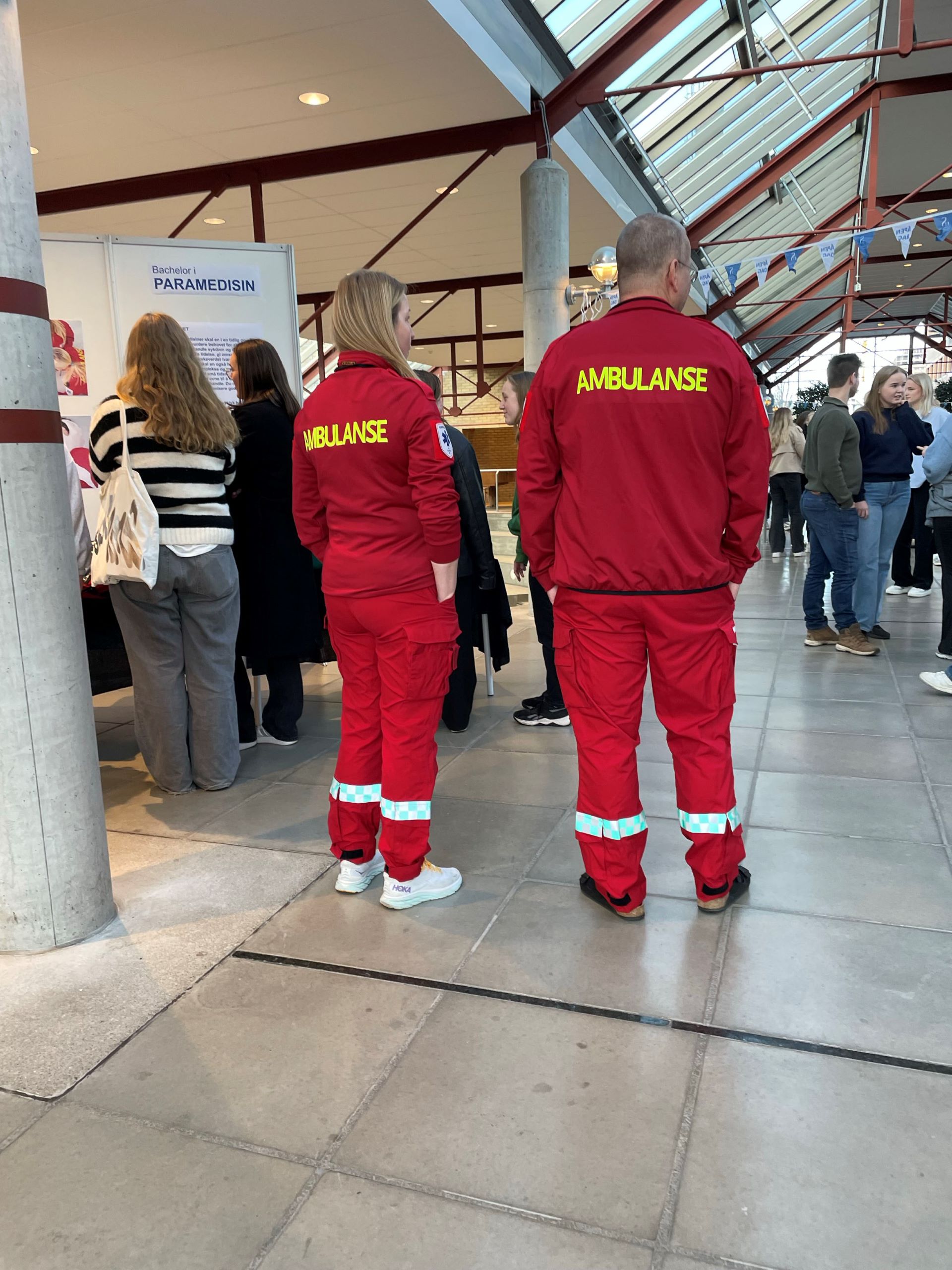 To personer i ambulanseuniform. Flere folk i bakgrunnen.