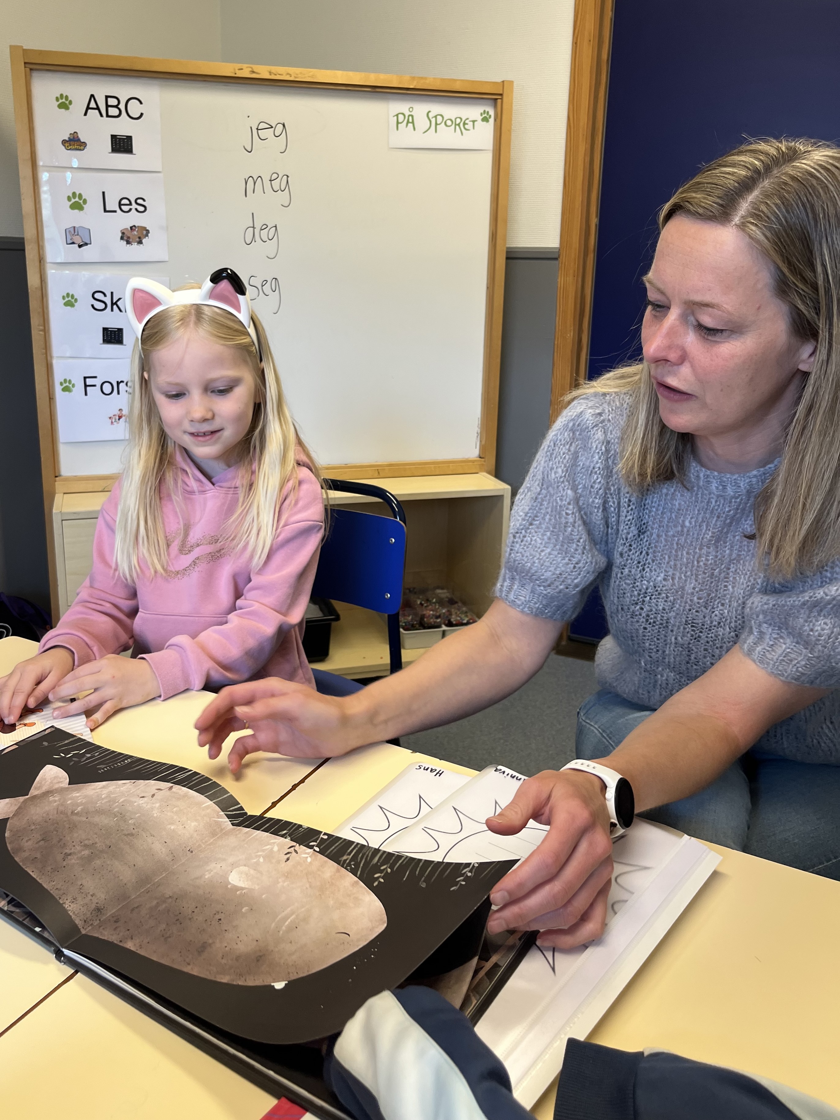 Jente og kvinne sitter i klasserom og ser i bok.