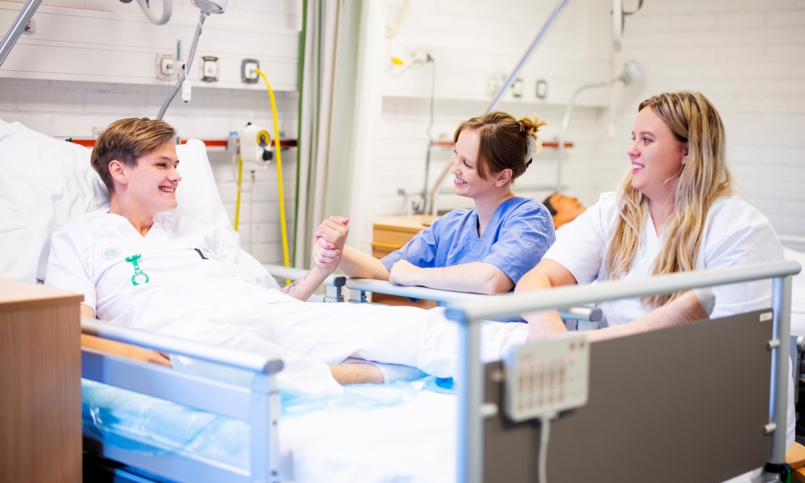 Tre sykepleierstudenter i treningsrom på campus. En mannlig student i hvit uniform spiller pasient og ligger i en pasientseng. To kvinnelige studenter sitter ved sengekanten og smiler til "pasienten". Den ene holder ham i hånden. 