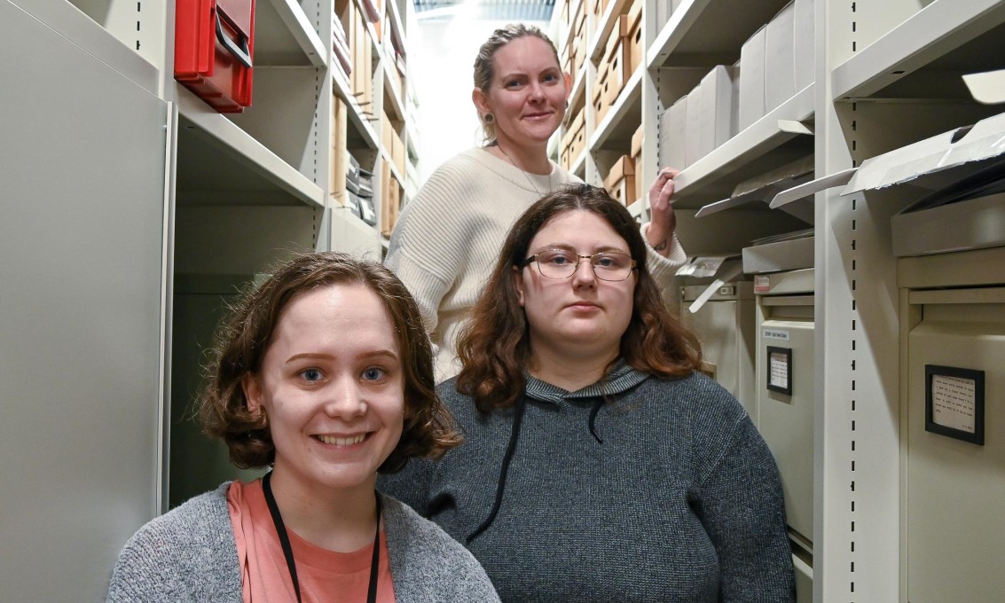 Tre unge kvinner står mellom to arkivhyller og ser rett i kamera