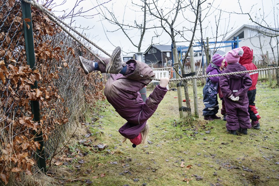 Fire barn står på en plen og snakker sammen. Hodene er tett sammen. Litt lenger unna turner ei jente opp ned etter beina i et tau.