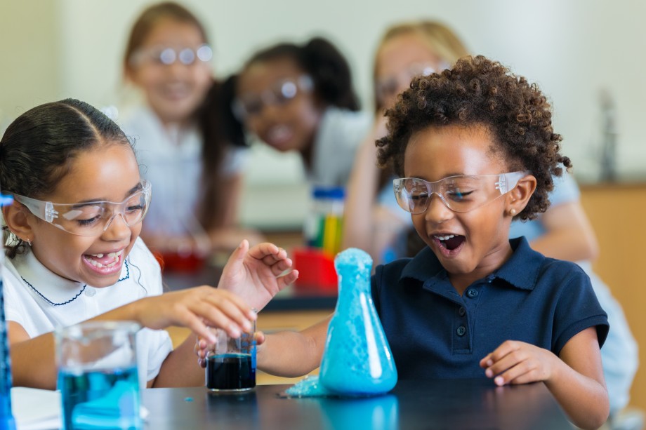 En gutt og en jente gjør et kjemisk eksperiment. De to har begge åpen, smilende munn og ser blå væske boble opp fra en glasskoble.