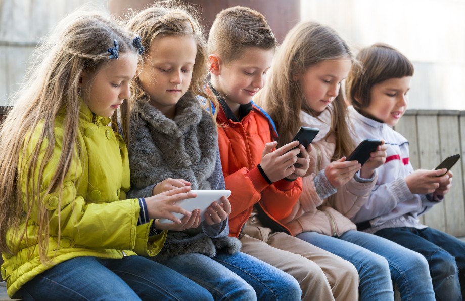 Fem barn sitter på rekke og rad og ser på mobil og nettbrett.